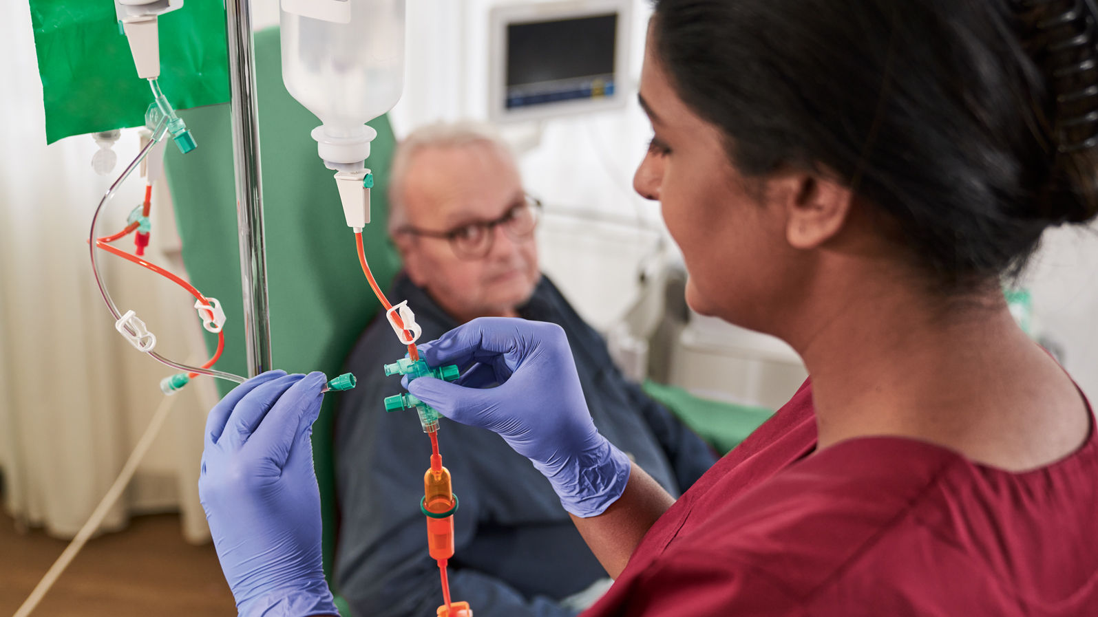 Onkologipatient med glasögon som sitter på en stol och sjuksköterska med handskar som hanterar cytoset 