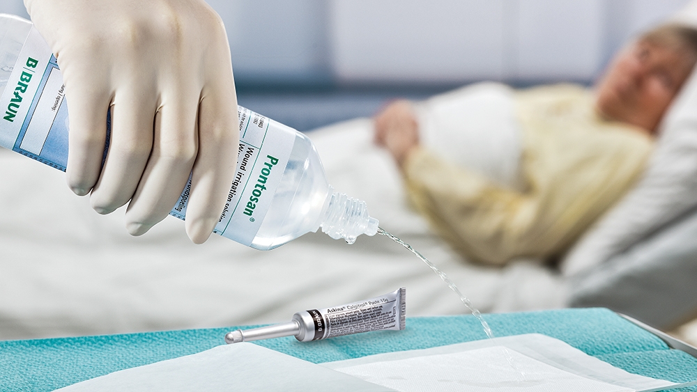 I förgrunden häller en hand med medicinsk handske Prontosan spolningslösning på en dyna som ligger på bordet bredvid ett Askina Calgitrol-rör. I bakgrunden ligger en kvinnlig patient i en sjukhussäng.