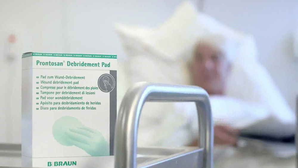 en förpackning Prontosan® Debridement Pad i fokus, med en patient liggande på en säng i bakgrunden