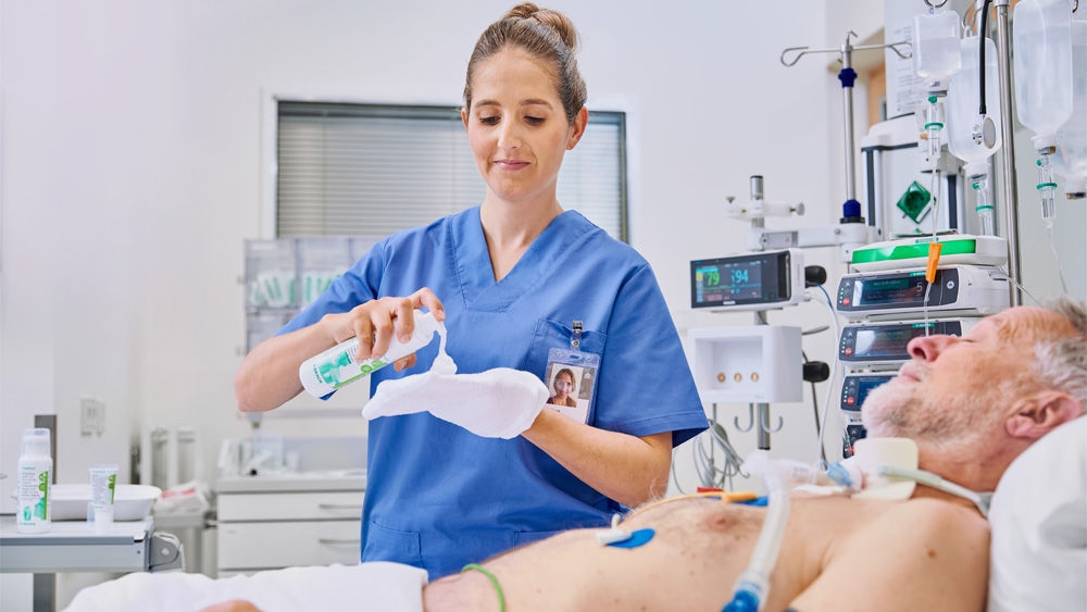 kvinnlig sjuksköterska som håller Prontoderm Foam på sjukhus framför en äldre patient som ligger i sängen och applicerar det med en handske