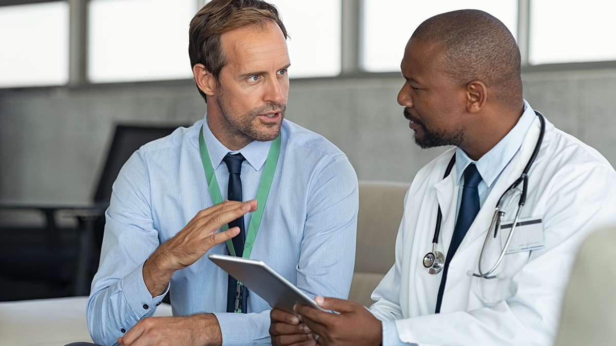 Läkemedelsrepresentant som pratar med läkare som sitter i en soffa. Afrikansk mogen läkare som diskuterar resultaten av analysen med specialist medan han konsulterar diagnosen på en digital surfplatta. Läkare i samtal med medicinsk rådgivare i sjukhusrum som diskuterar patientrapportärende.