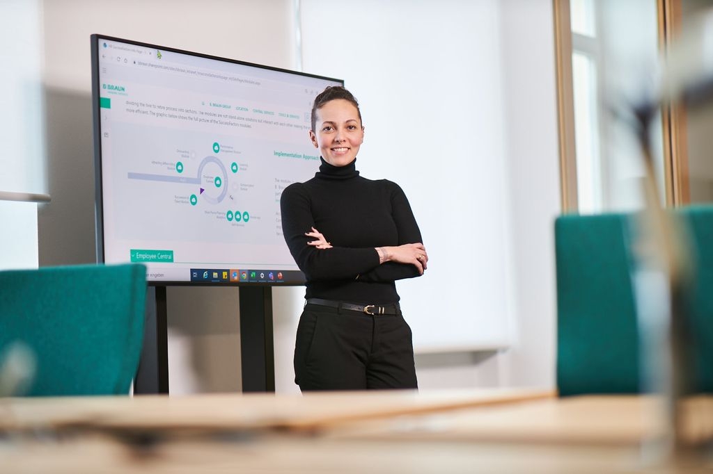 B. Renata Almeida, Braun-anställd, står och ler framför en TV som visar grafer på ett kontor. 