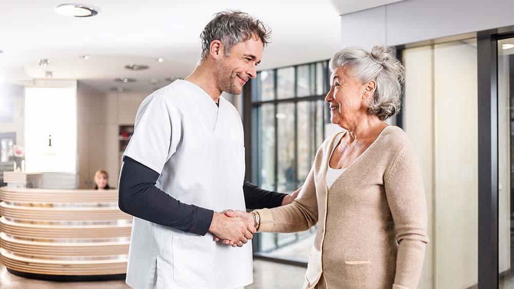 En läkare skakar hand med en patient, i bakgrunden finns en reception