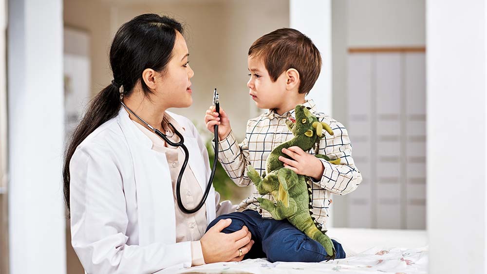 En kvinnlig vårdgivare pratar med en liten pojke som håller en leksaksdinosaurie. Pojken ser engagerad och nyfiken ut, medan vårdgivaren ser på honom varmt och vänligt.