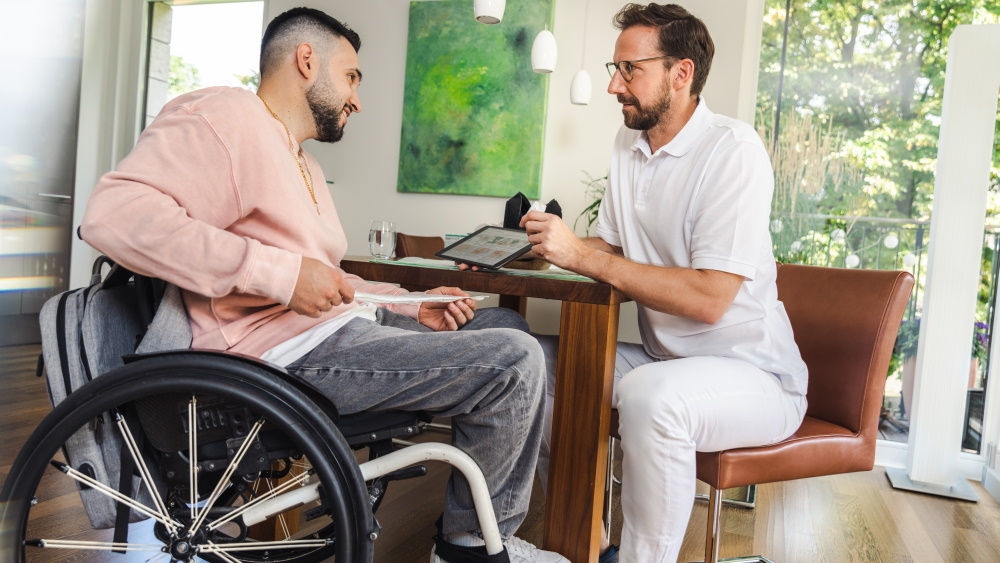 En man sitter i rullstol och håller en kateter i händerna. Framför honom sitter en hälsospecialist på ett sjukhus och ger instruktioner med hjälp av en surfplatta