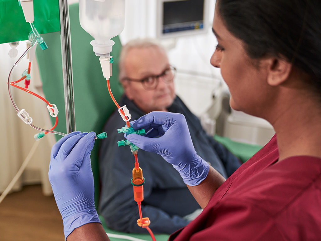 Onkologipatient med glasögon sitter på en stol och sjuksköterska med handskar hanterar CytoSet 