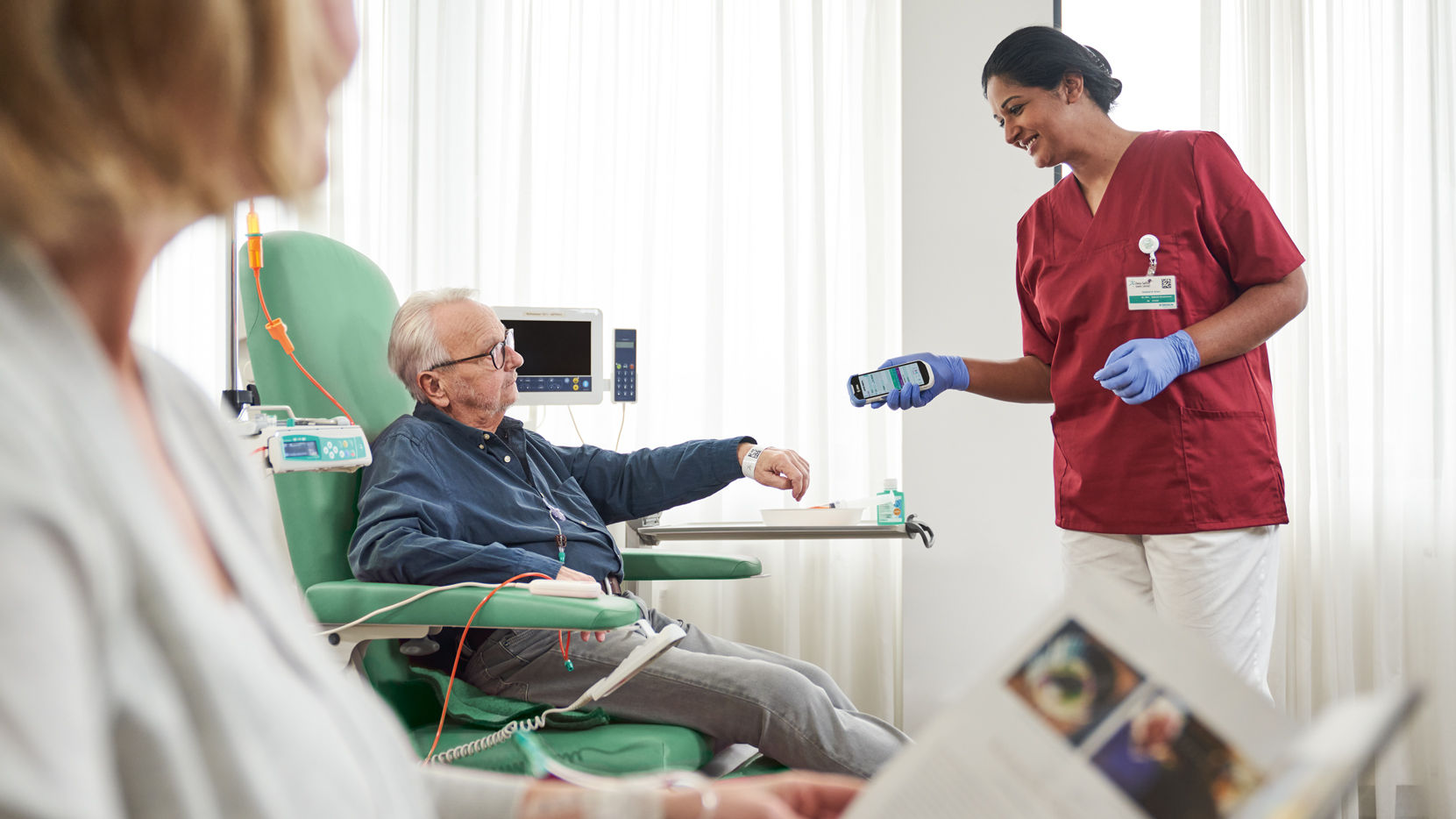 Onkologipatient med glasögon sitter på en stol och en sjuksköterska med handskar skannar patient-ID på handledsbandet för säkerhet och dokumentation 