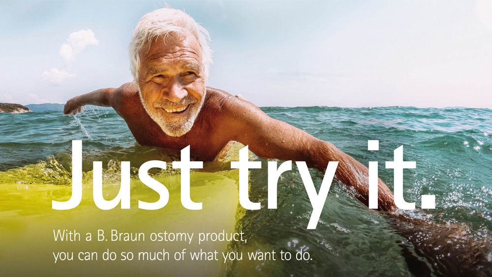 En äldre man med vitt hår ligger på en gul surfbräda och paddlar i havet under en klarblå himmel.