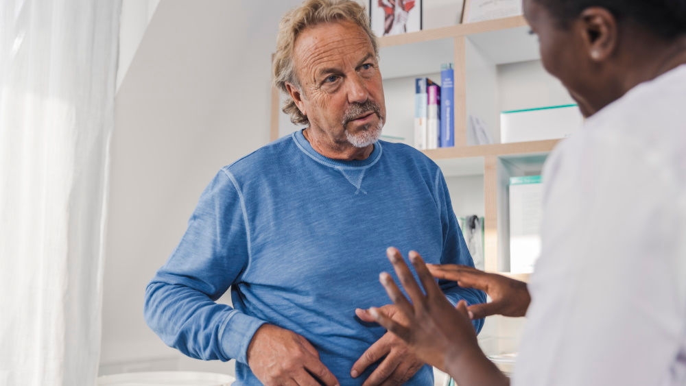 en medelålders man ställer en fråga till läkaren medan han håller i höger sida av magen, en manlig läkare förklarar något för honom