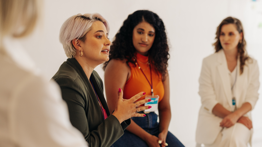 Fyra kvinnor sitter sida vid sida i Women's Mentoring Program. Målet är att öka kvinnors synlighet i organisationen och hjälpa dem i deras personliga utveckling. En andra kvinna tittar på den första, en gråhårig kvinna i grön jacka, medan hon pratar och ger en handsignal.  