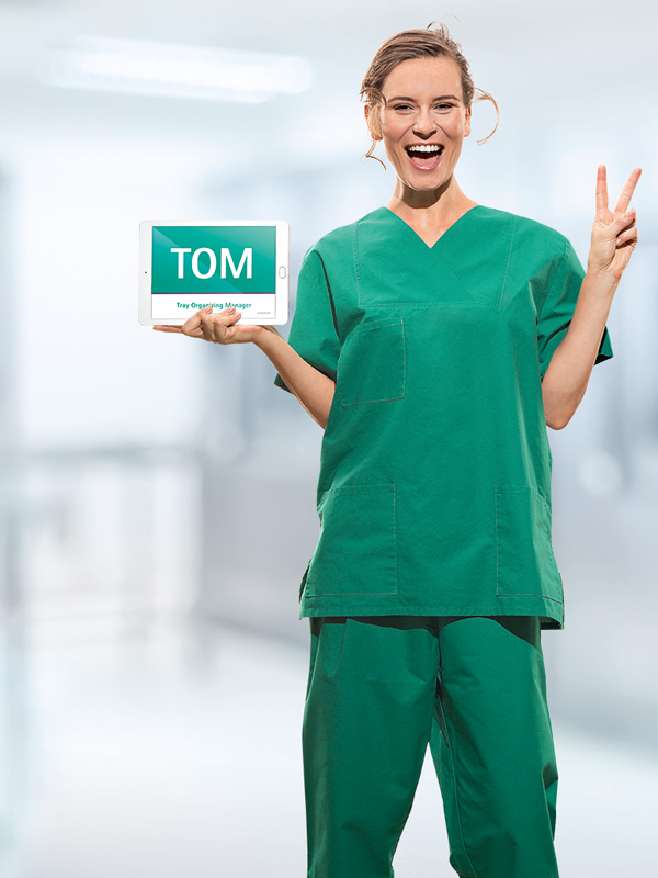 En kvinnlig läkare i medicinska kläder håller en läsplatta med förkortningen ”TOM” på. Hon visar också ett ”V”-tecken med fingrarna