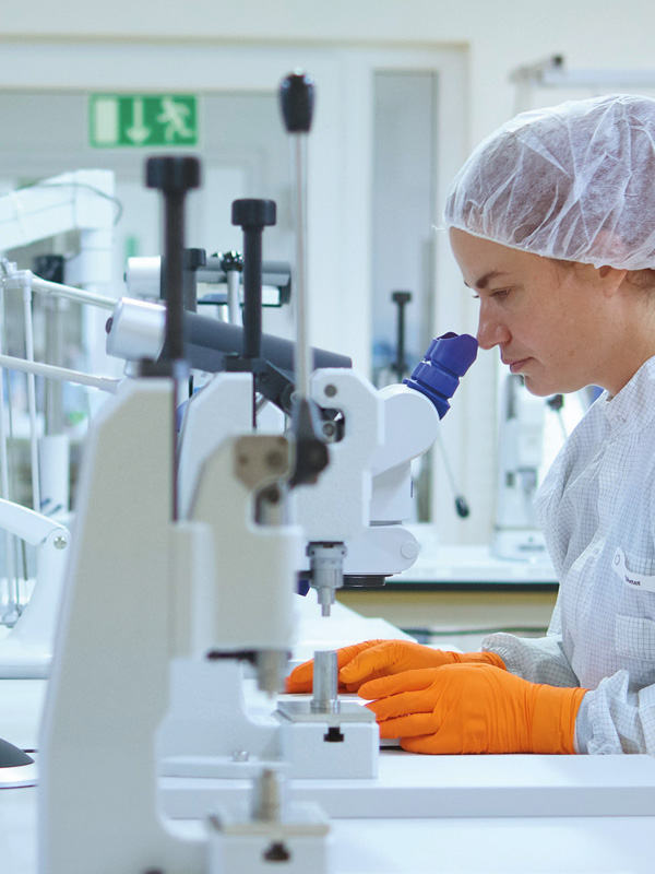 En kvinnlig vårdpersonal sitter vid ett bord och analyserar prover genom ett medicinskt mikroskop