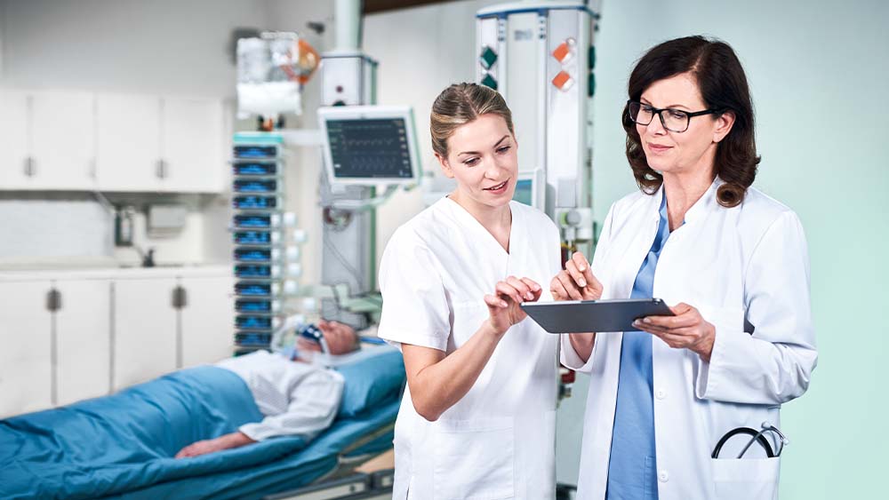 En läkare och en sjuksköterska diskuterar doseringen för B. Braun smart infusion, som matas in direkt från en surfplatta. Patienten på intensivvårdsavdelningen kan ses i bakgrunden, ansluten till en ställning full av smarta infusionsenheter.
