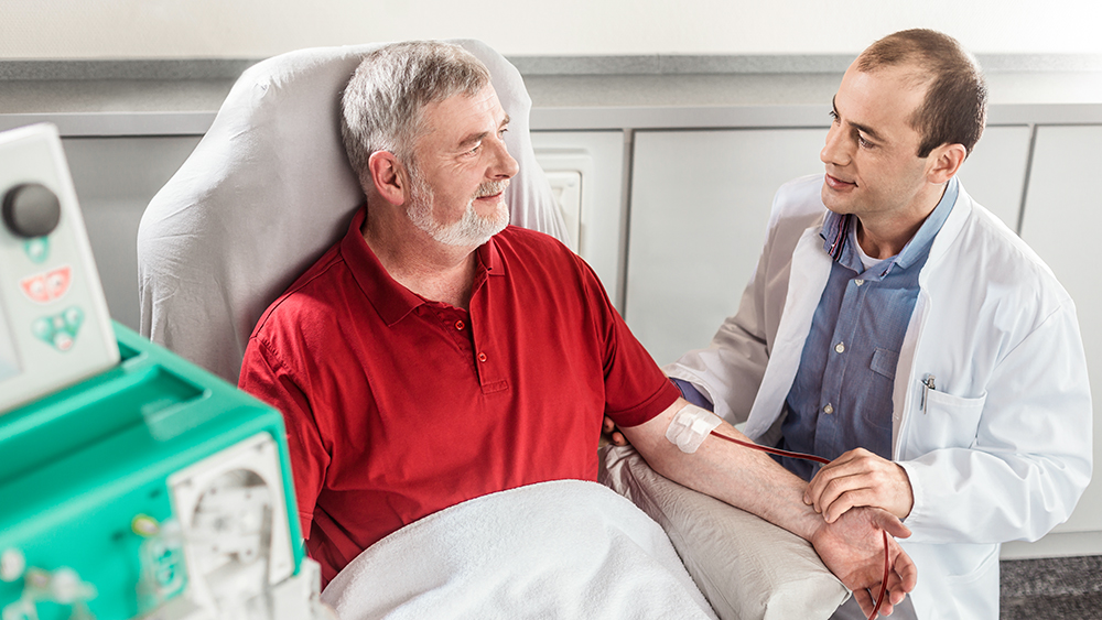 En patient sitter i en stol med en filt över sig bredvid en dialysmaskin. Läkaren sitter bredvid patienten.