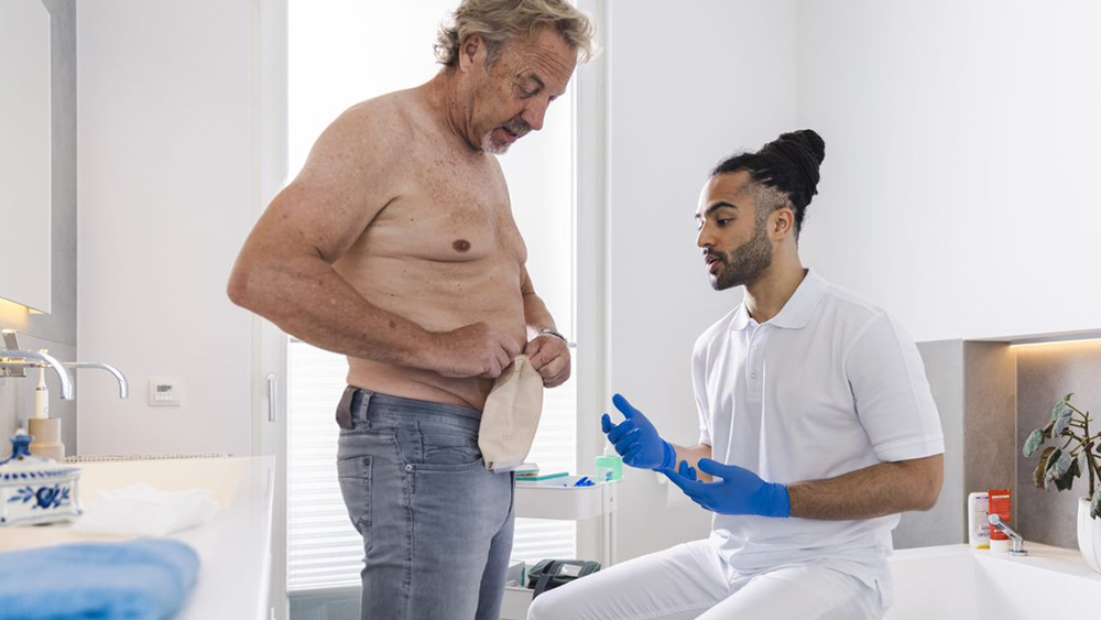 Man med bar överkropp i privat badrum rör stomipåse på magen. Manlig sjuksköterska sitter på badkarskanten och ger instruktioner.