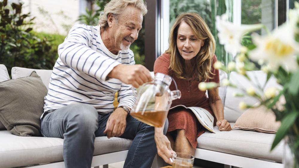 En medelålders man och en medelålders kvinna sitter tillsammans i soffan och mannen häller upp en kopp te till kvinnan