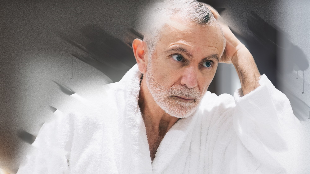 En äldre patient i badrock tittar på sig själv i badrumsspegeln.